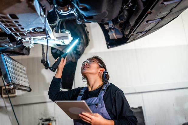 Female mechanic using flashlight and digital tablet, inspecting underside of car raised on lift in auto repair shop