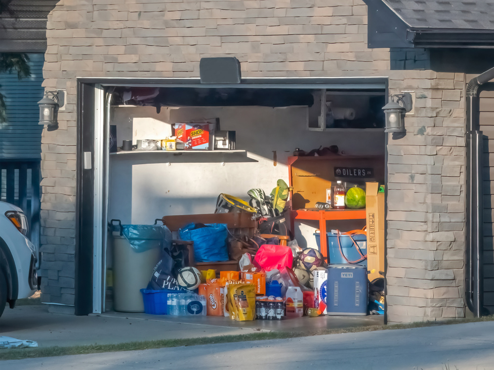 Calgary, Alberta, Canada. Jun 8, 2025. A sunlit garage stands open, revealing a clutter of household items, storage bins, hinting at a need for organization