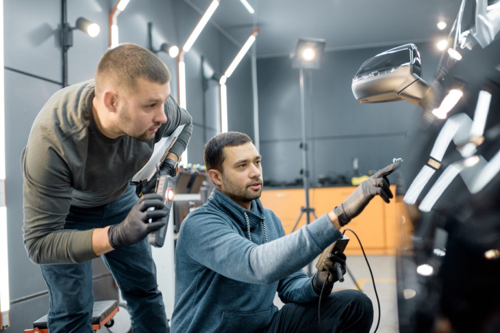 Two car service workers examining vehicle body for scratches and damages while taking a car for professional automotive detailing