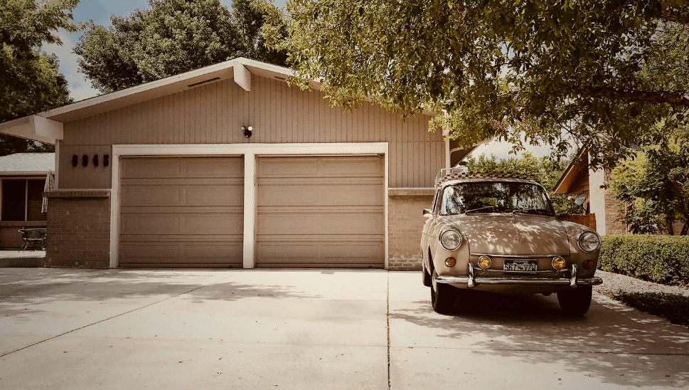 A picture of an old car parked on a driveway leading up to a double-door garage.